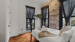 A modern hotel room with a brick wall, large windows with sheer curtains, a wooden floor, and a neatly made bed with a towel and a blanket.