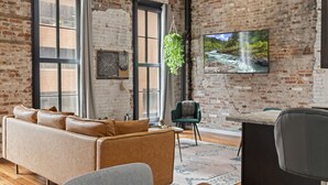 A modern living room with exposed brick walls, a leather sofa, a green armchair, and a large TV mounted on the wall.