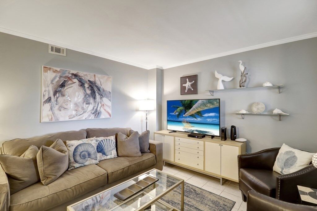 Ein Wohnzimmer mit einem beigen Sofa, einem Glastisch, einem Flachbildfernseher und Dekorationen auf Regalen.