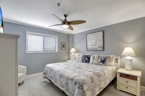 A bedroom with a bed, a ceiling fan, a window with blinds, a nightstand with a lamp, and a wall-mounted picture. - Escaptopia - Mid Island Villa with Multiple Pools by RedAwning (Hilton Head Island)