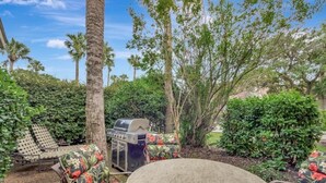 A patio with a round table, floral chairs, a grill, and surrounding greenery. - Turtle's Tide - Spacious Villa in Queens Grant with Private Patio by RedAwning (Hilton Head Island)