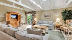 A spacious living room with a large sofa, a television, and a dining table. There is a window with a view of greenery outside. - Turtle's Tide - Spacious Villa in Queens Grant with Private Patio by RedAwning (Hilton Head Island)