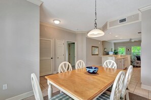 A dining room with a wooden table and white chairs, a kitchen in the background, and a ceiling light.