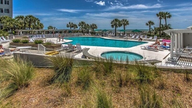 A swimming pool with lounge chairs and palm trees, surrounded by a building. - Shorenugh - Bright Villa with Rooftop Pool Next to Coligny Plaza by RedAwning (Hilton Head Island)