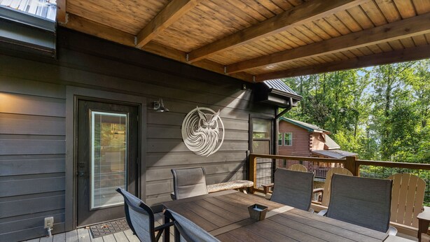 A covered outdoor deck with a wooden table and chairs, a metal owl decoration, and a view of a forested area. - Night Owl 3 Bedroom Cabin by RedAwning (Gatlinburg)