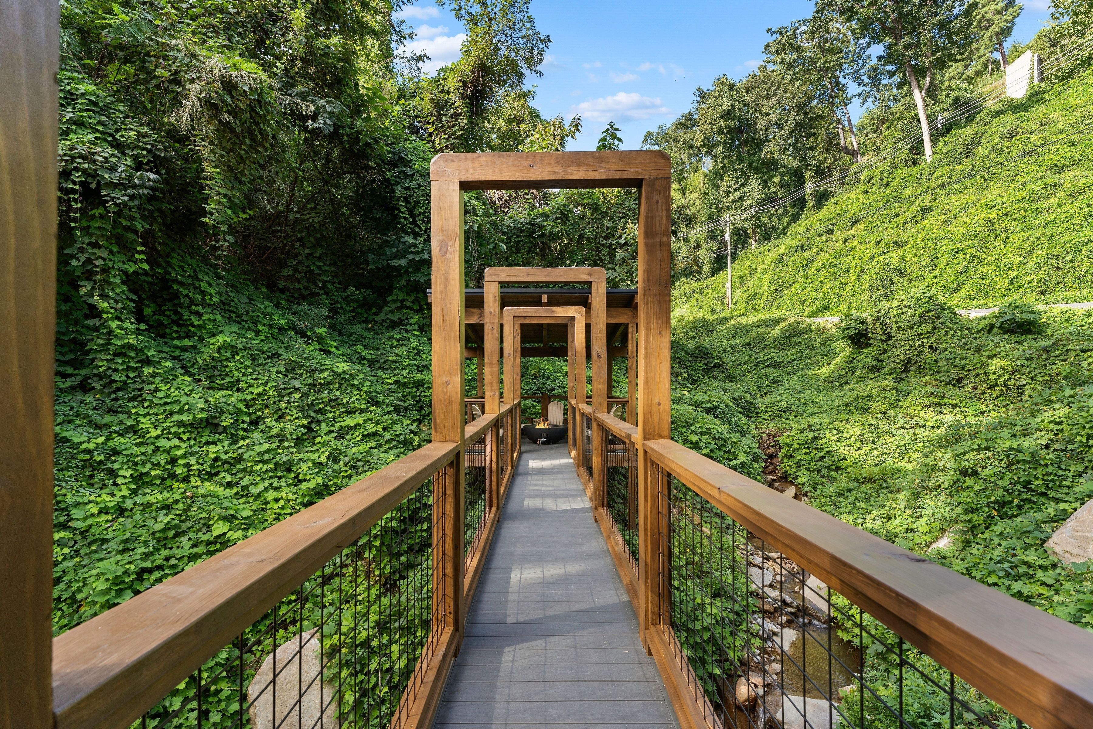 A wooden pedestrian bridge with a metal railing, surrounded by lush greenery and a small stream.