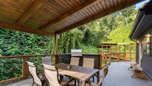 A covered outdoor deck with a wooden table and chairs, a barbecue grill, and a view of a lush green forest. - Night Owl 3 Bedroom Cabin by RedAwning (Gatlinburg)