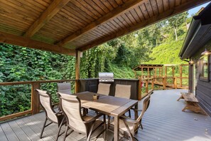 A covered outdoor deck with a wooden table and chairs, a barbecue grill, and a view of a lush green forest. - Night Owl 3 Bedroom Cabin by RedAwning (Gatlinburg)