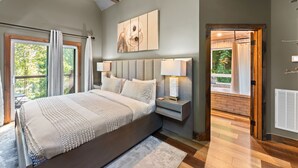 A bedroom with a large bed, a nightstand, a wall-mounted lamp, and a view of the outdoors through a window. - Night Owl 3 Bedroom Cabin by RedAwning (Gatlinburg)