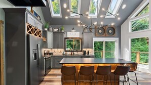 A modern kitchen with a central island, bar stools, and a view of greenery through large windows. - Night Owl 3 Bedroom Cabin by RedAwning (Gatlinburg)