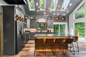 A modern kitchen with a central island, bar stools, and a view of greenery through large windows. - Night Owl 3 Bedroom Cabin by RedAwning (Gatlinburg)