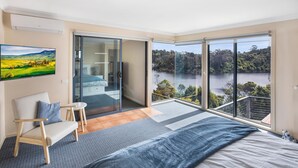 Ein Schlafzimmer mit einem großen Bett, einem Sessel, einem kleinen Tisch und einer Glasraumtür, die auf einen Balkon mit Seeblick führt.