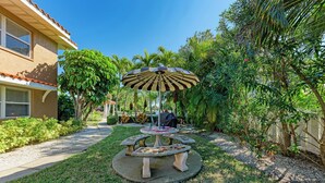 A circular outdoor seating area with a central table and a large umbrella, surrounded by a well-maintained garden with various plants and a pathway leading to a building. - Casa Sierra 203B - 2BR Condo with Pool - By Island Real Estate Vacations (Holmes Beach)