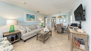 A living room with a sofa, coffee table, and TV. There are bookshelves and a lamp. - Casa Sierra 203B - 2BR Condo with Pool - By Island Real Estate Vacations (Holmes Beach)