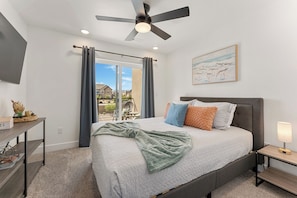 A bedroom with a bed, a ceiling fan, a bedside table with a lamp, a window with curtains, and a painting on the wall.