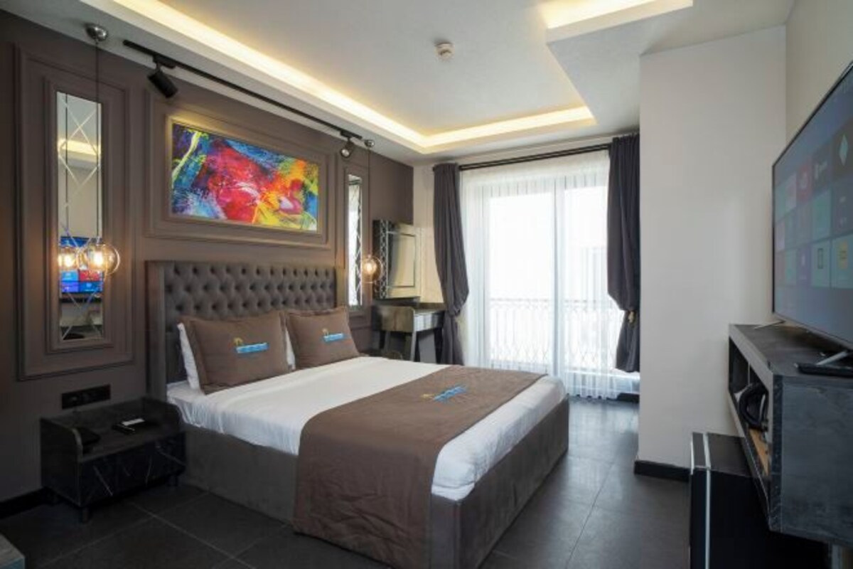 A modern hotel room with a large bed, a flat-screen TV, a desk, and a window with curtains.