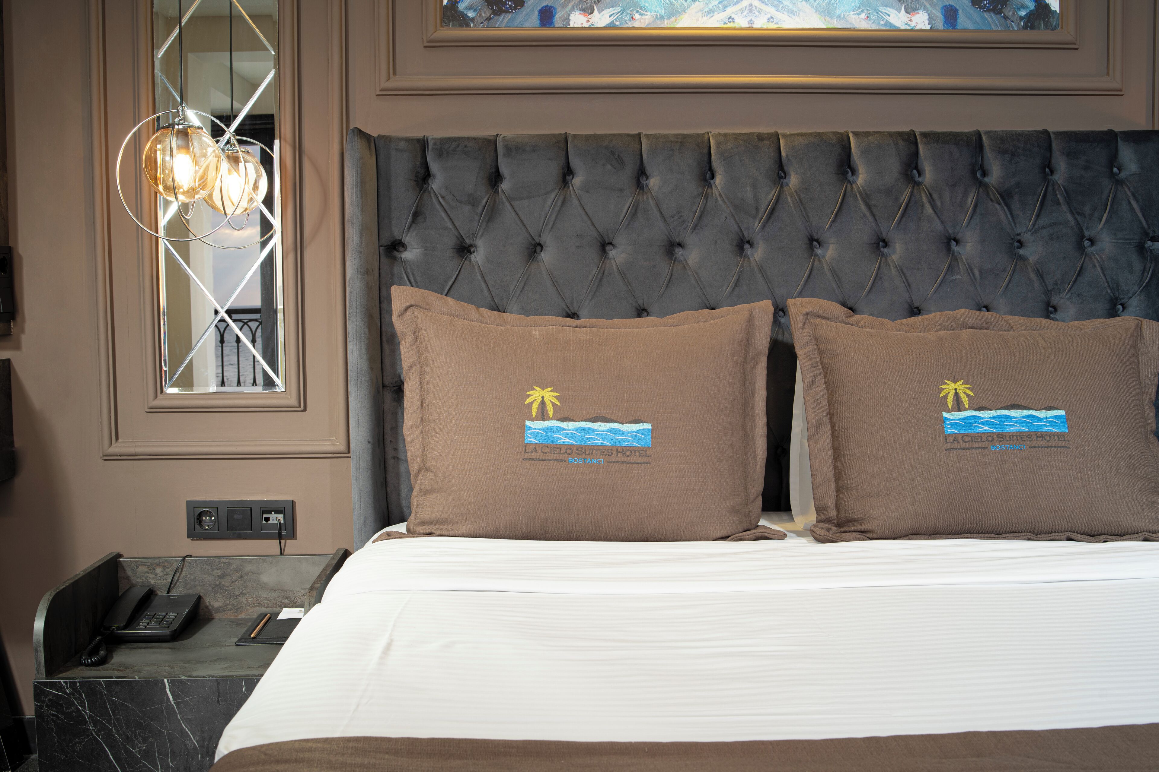 Una habitación de hotel con un cabecero capitonado, dos almohadas con diseño tropical, un teléfono y una mesita de noche con una lámpara.
