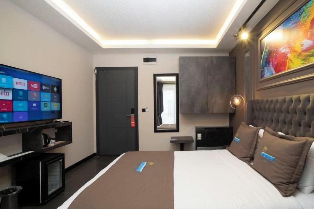 A hotel room with a large flat-screen TV displaying streaming services, a bed with brown bedding, a bedside table, a small cabinet, and a large window with a view.