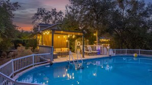 A swimming pool with a covered seating area, lounge chairs, and string lights.