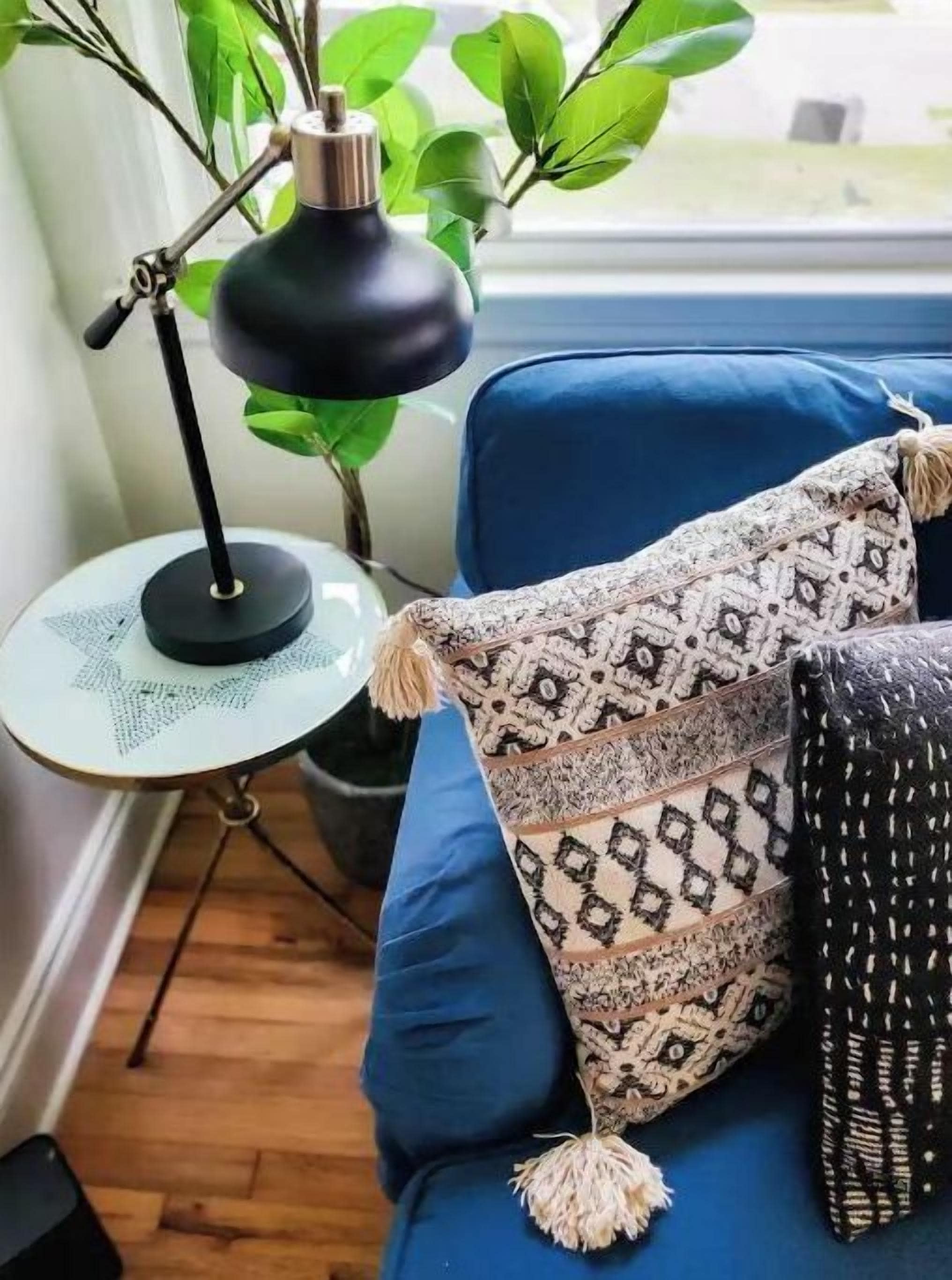 Een blauwe bank met gestreepte kussens en een franje, een klein rond tafeltje met een lamp en een bloempot.