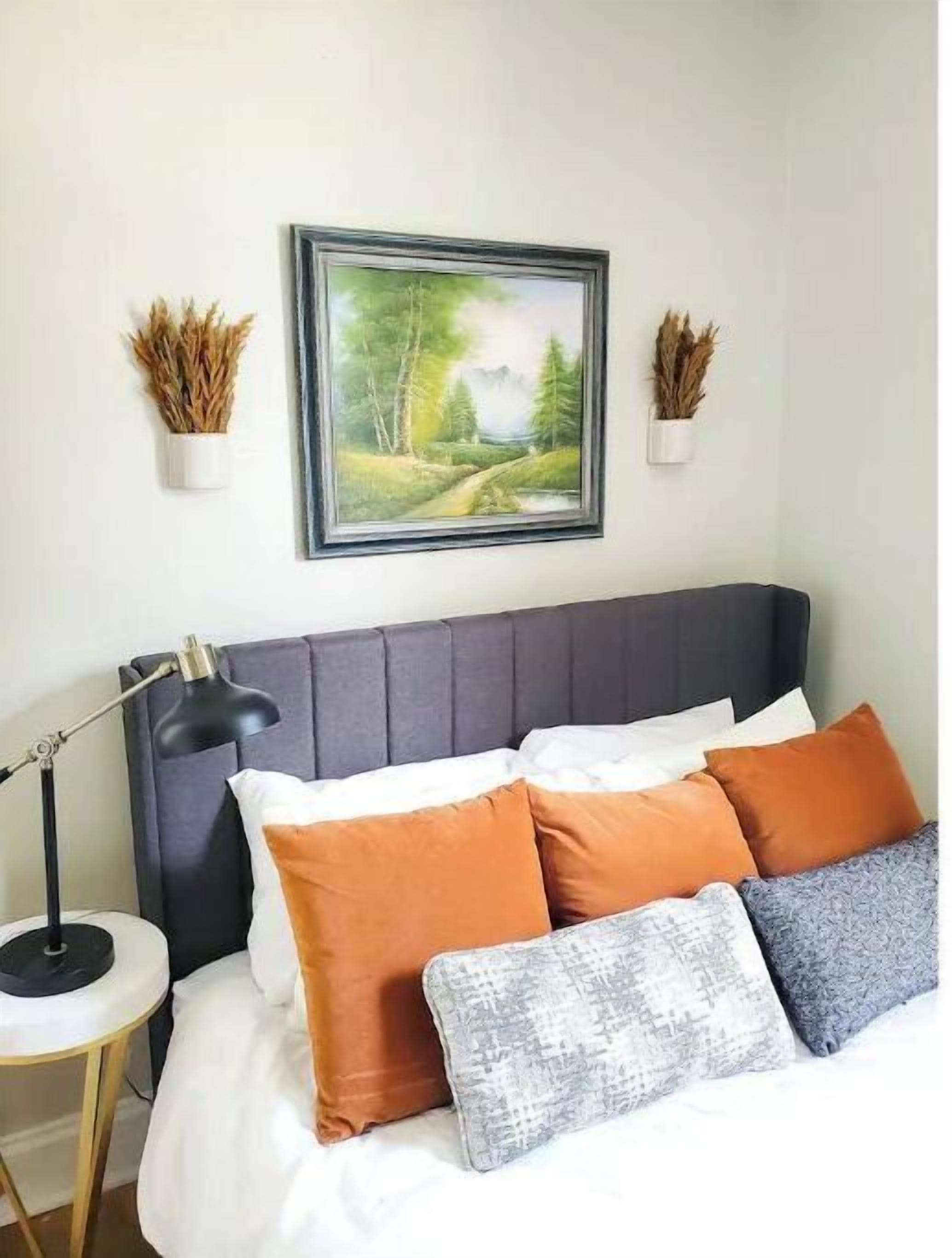 Un dormitorio con cama, almohadas, una mesita de noche, una lámpara y un cuadro en la pared.