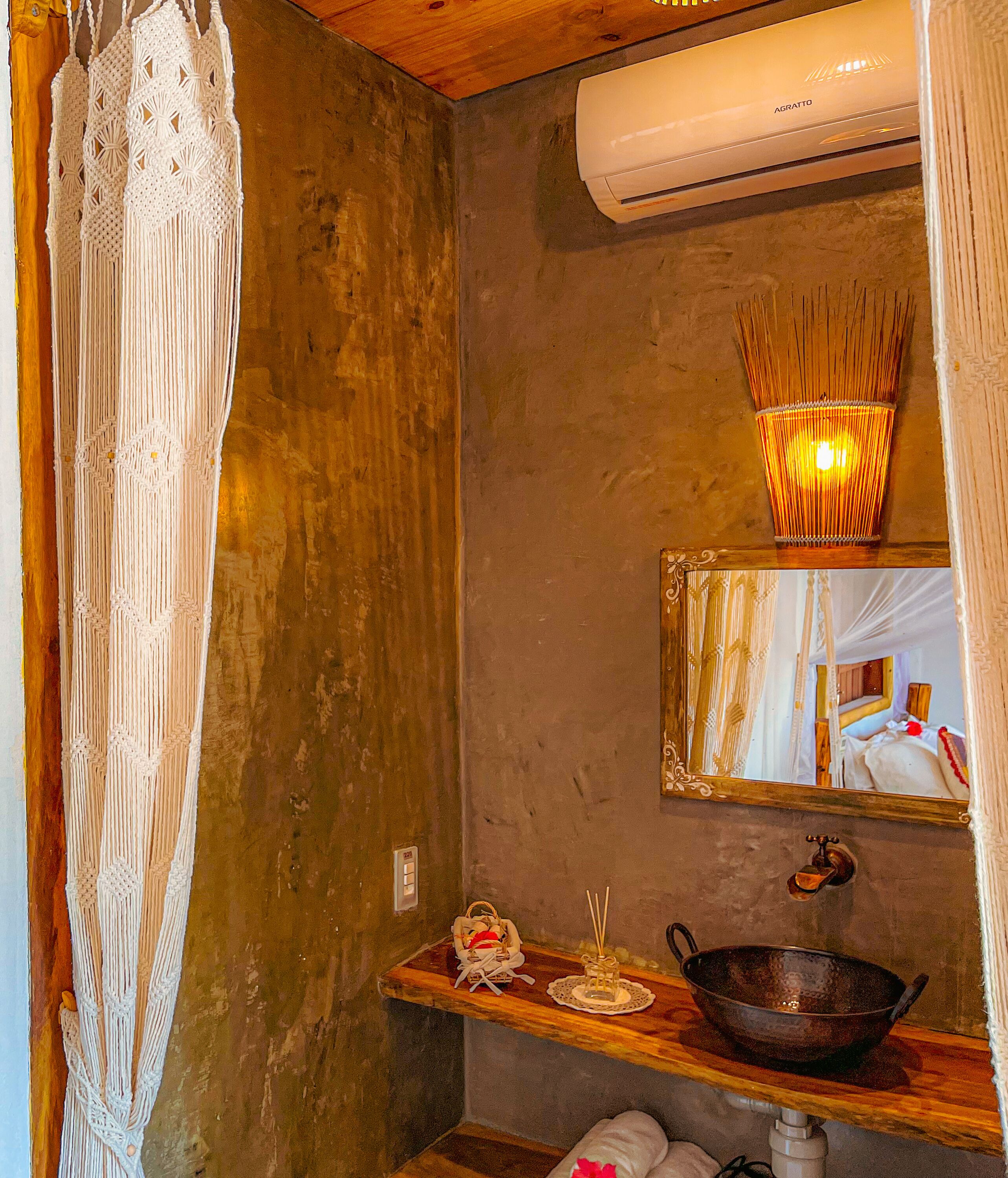 A bathroom with a macrame curtain, a mirror, a sink with a bowl, and an air conditioner.