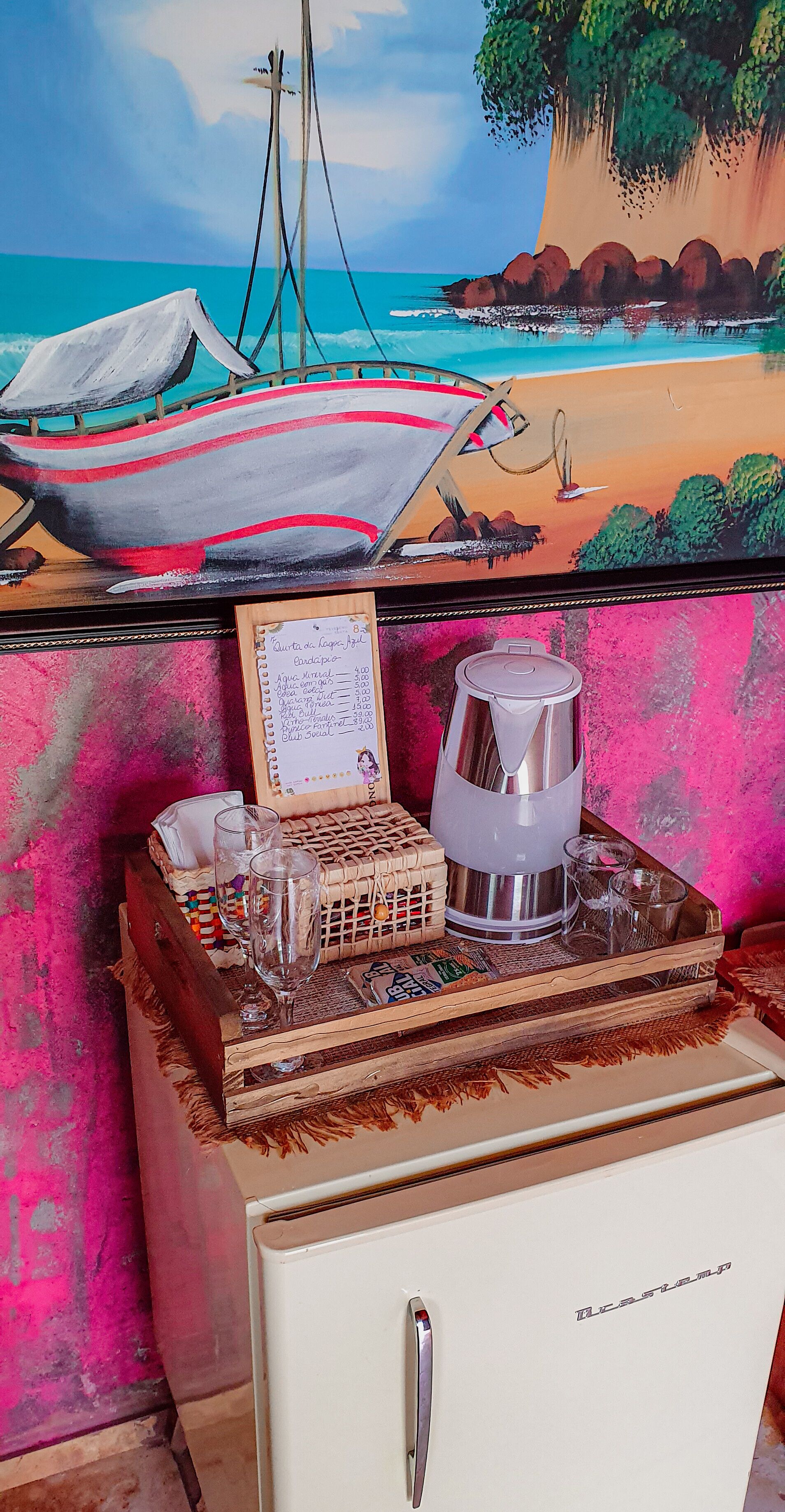 Una habitación con un cuadro de una escena de playa, una bandeja de madera con vasos y un menú, y un refrigerador.