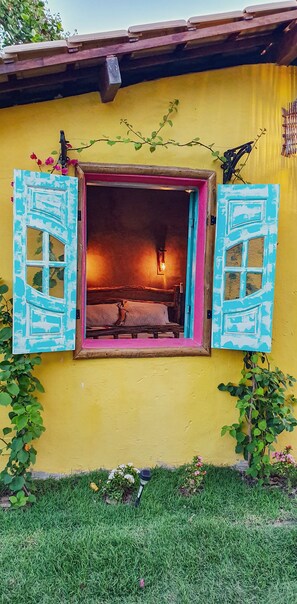 A yellow building with a window featuring blue shutters, a bed inside, and a small plant on the windowsill.