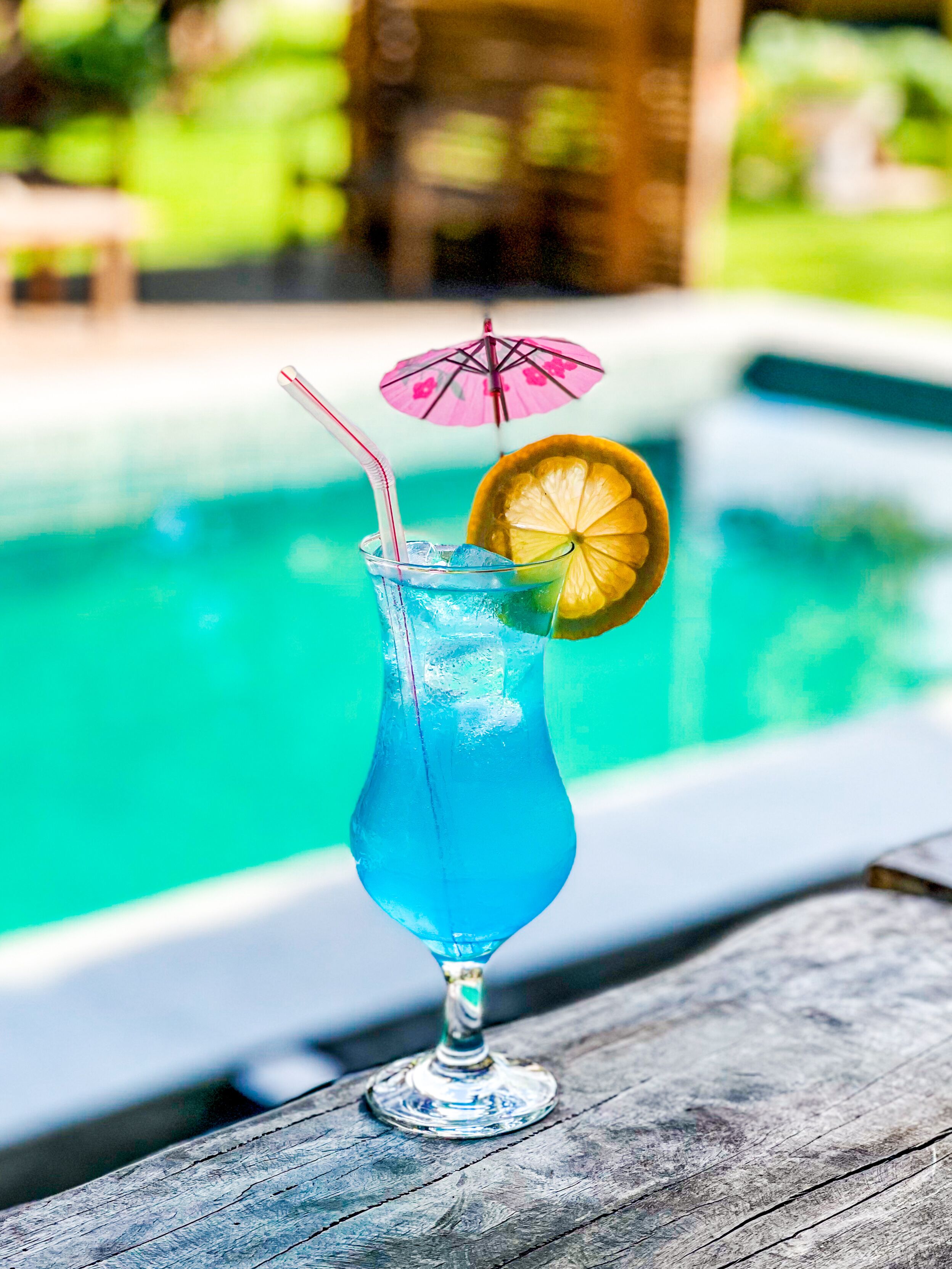A cocktail with a straw and a slice of lemon in a tall glass, placed on a wooden surface near a swimming pool.