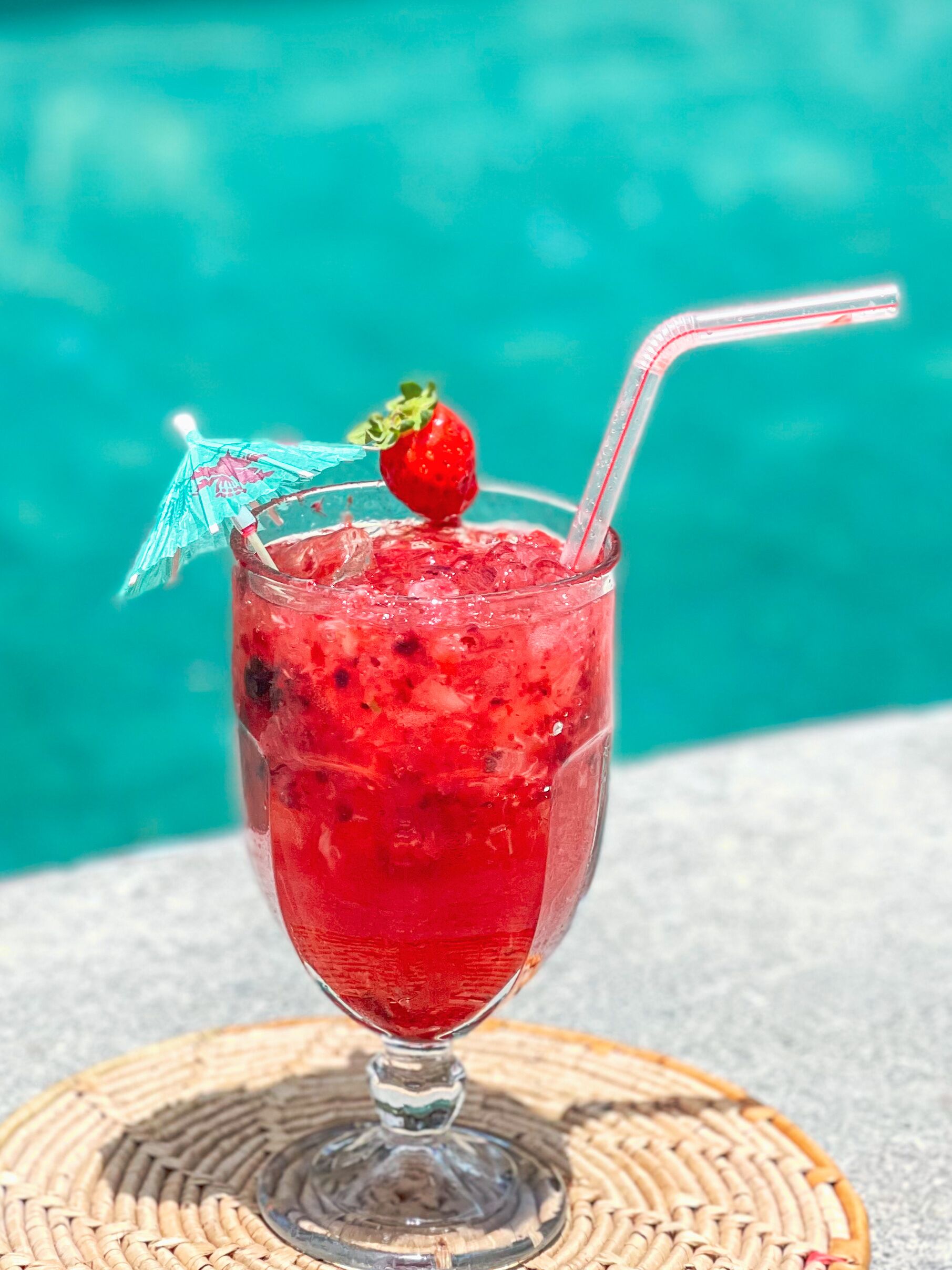 Ein Cocktail mit Erdbeere und einem gestreiften Strohhalm, platziert auf einer gewebten Unterlage am Pool.