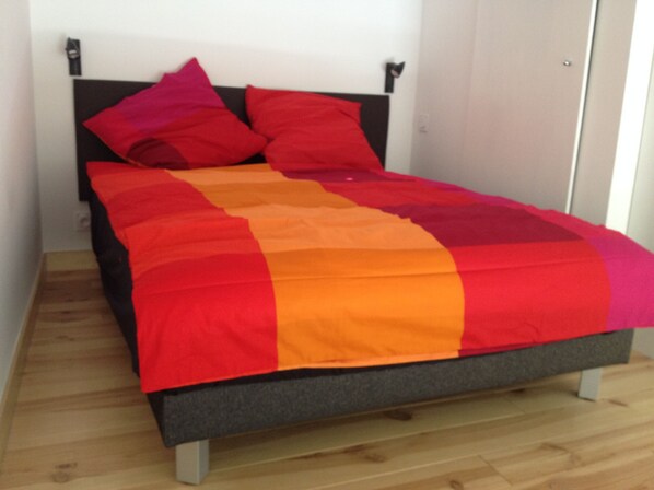 A bed with a multicolored blanket and two pillows, a wooden floor, and a white wall with a door.