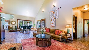 A spacious living room with a high ceiling, a dining area with a wooden table and chairs, and a large window providing natural light. - Dancing Ferns in Volcano, Hawaii (Volcano)
