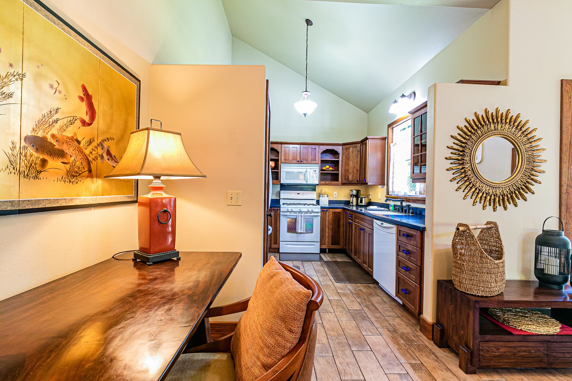Cocina con armarios de madera, estufa y microondas. Hay una mesa de comedor con sillas y una lámpara.