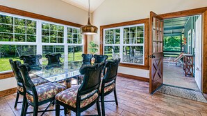 A dining area with a glass table and chairs, large windows, and a wooden door leading outside. - Dancing Ferns in Volcano, Hawaii (Volcano)