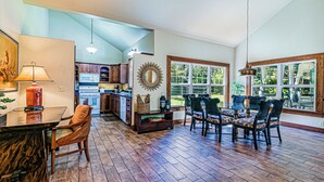A spacious kitchen with wooden cabinets, a dining area with a table and chairs, and large windows offering views of the outdoors. - Dancing Ferns in Volcano, Hawaii (Volcano)