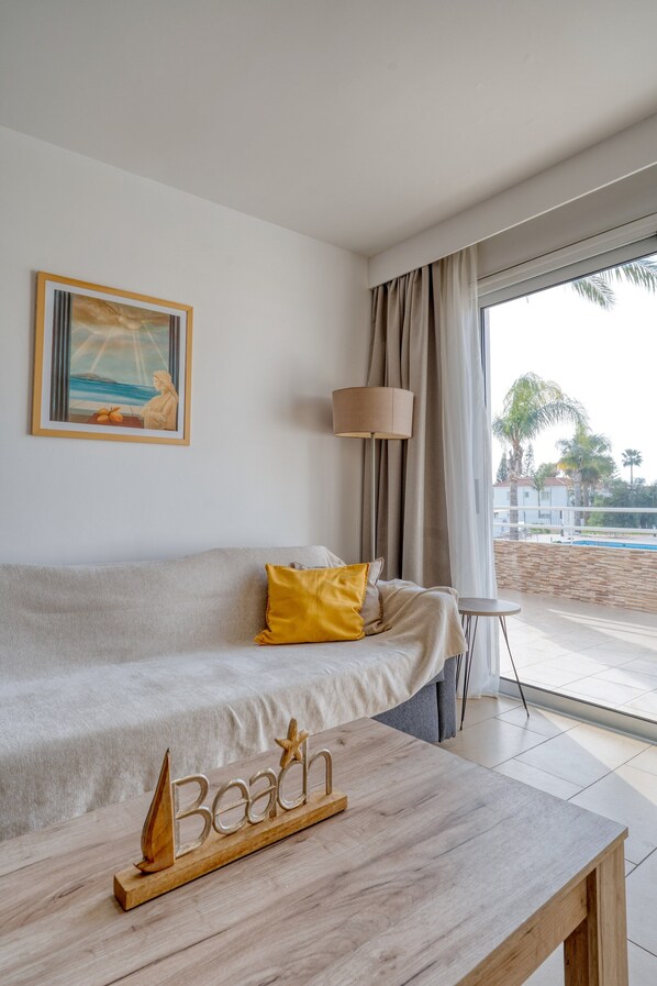 A modern room with a sofa, a coffee table, and a view of the beach through large windows. - Narcissos 'Nissi Beach' apartment C3 (Ayia Napa)