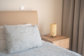 A neatly made bed with a light blue textured pillow and a matching blanket, a wooden bedside table with a woven lamp, and a plain wall with a light switch. - Narcissos 'Nissi Beach' apartment C3 (Ayia Napa)