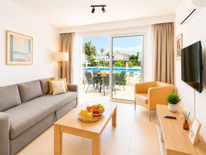 A modern hotel room with a sofa, a coffee table with fruit, a TV, and a balcony with a pool view.