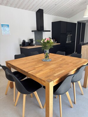 A dining area with a wooden table and black chairs, a vase with flowers, and a kitchen in the background. - House, villa with a year-round heated indoor pool set at 31° (St jean de monts)