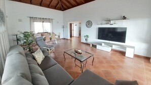 Un salon spacieux avec un plafond haut, un grand téléviseur à écran plat, un canapé et un coin repas comprenant des chaises et une table.