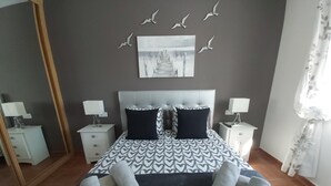 Une chambre à coucher bien aménagée, avec un grand lit, deux tables de chevet avec des lampes, un miroir et une œuvre d’art représentant des oiseaux en vol, accrochée au mur.