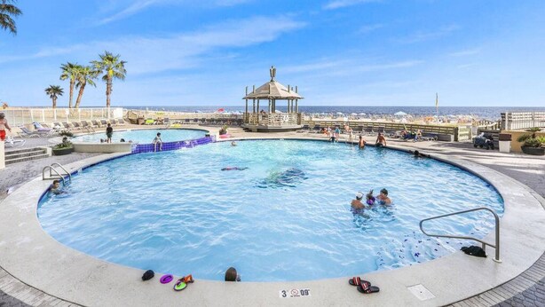 A large outdoor swimming pool with a gazebo, palm trees, and a view of the ocean.