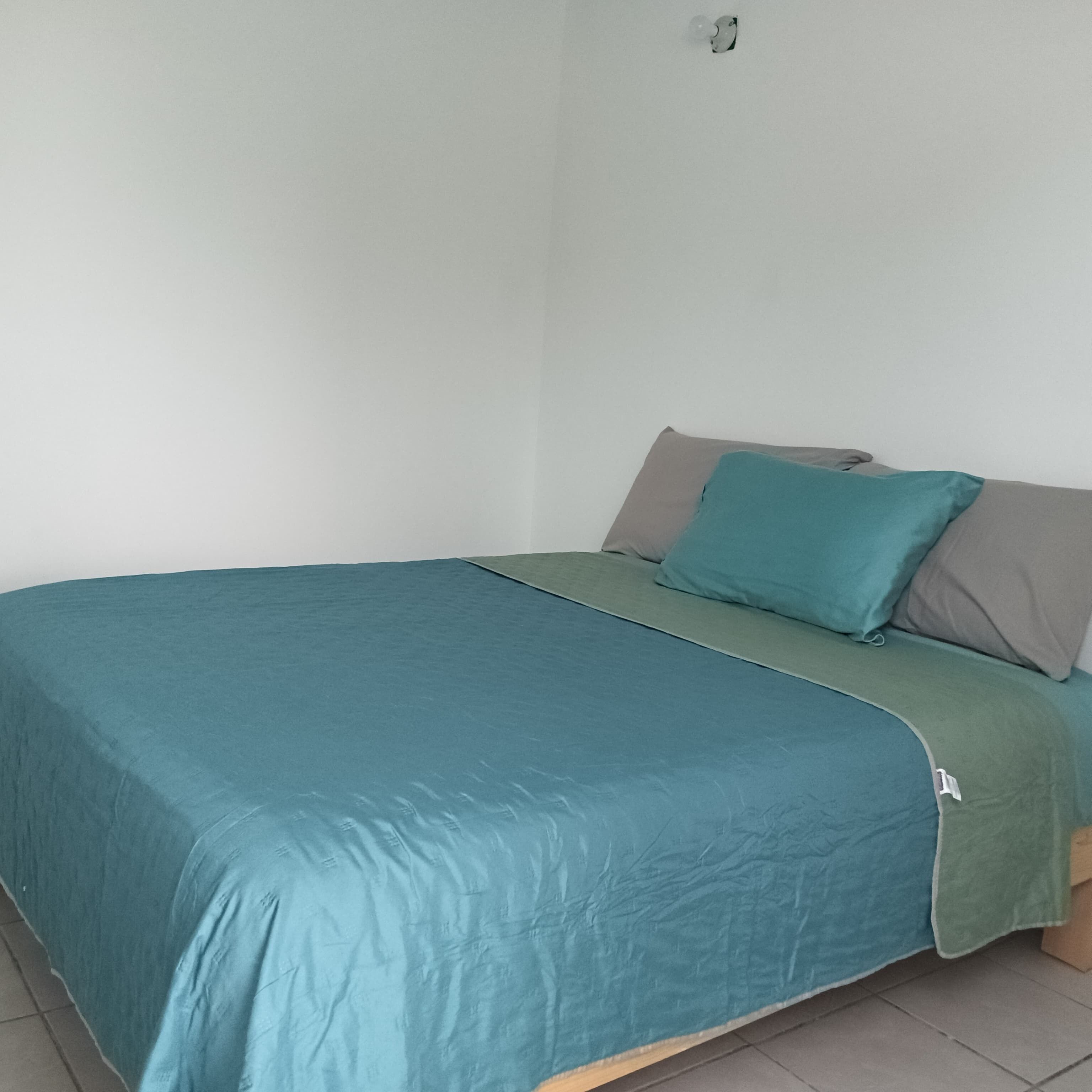 A single bed with a teal bedspread and two pillows, one teal and one gray, in a room with white walls and a tiled floor.