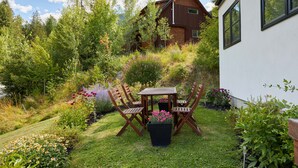 Une maison moderne dotée d’un toit en pente, entourée d’un jardin luxuriant agrémenté d’une table et de chaises en bois, ainsi que de diverses plantes et fleurs.
