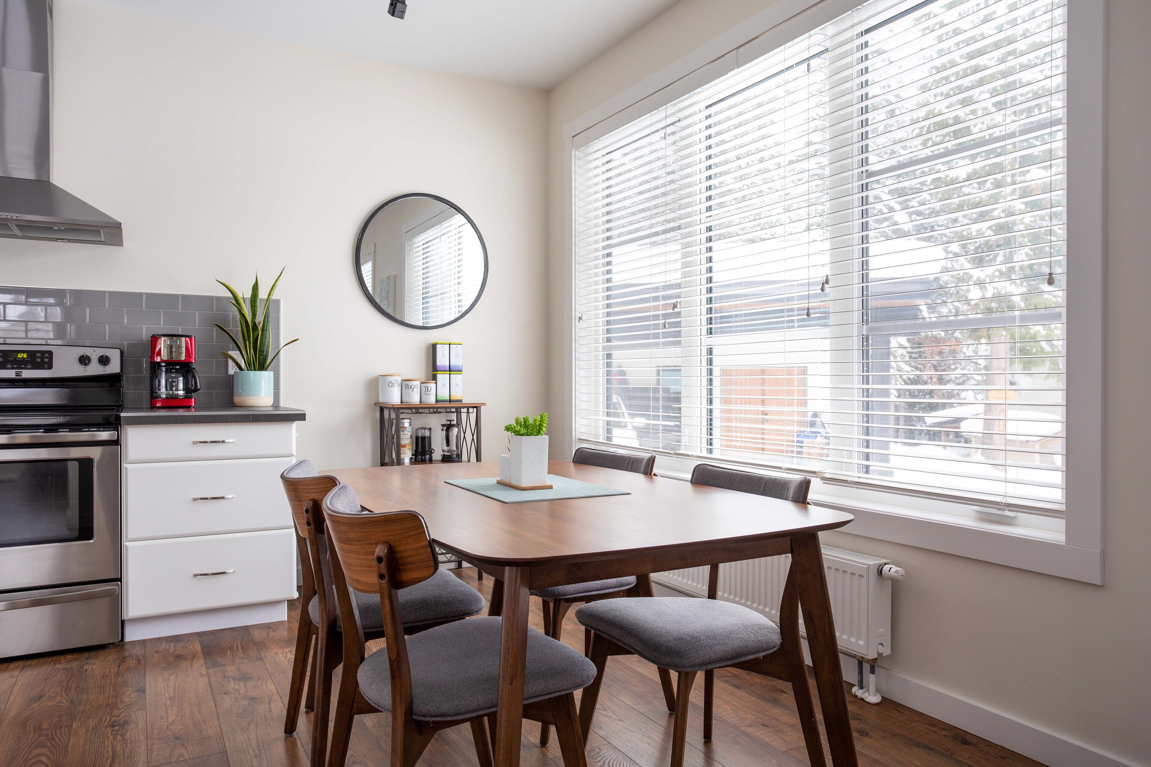 A modern kitchen with a dining area, a large window with blinds, and a view of the cityscape.