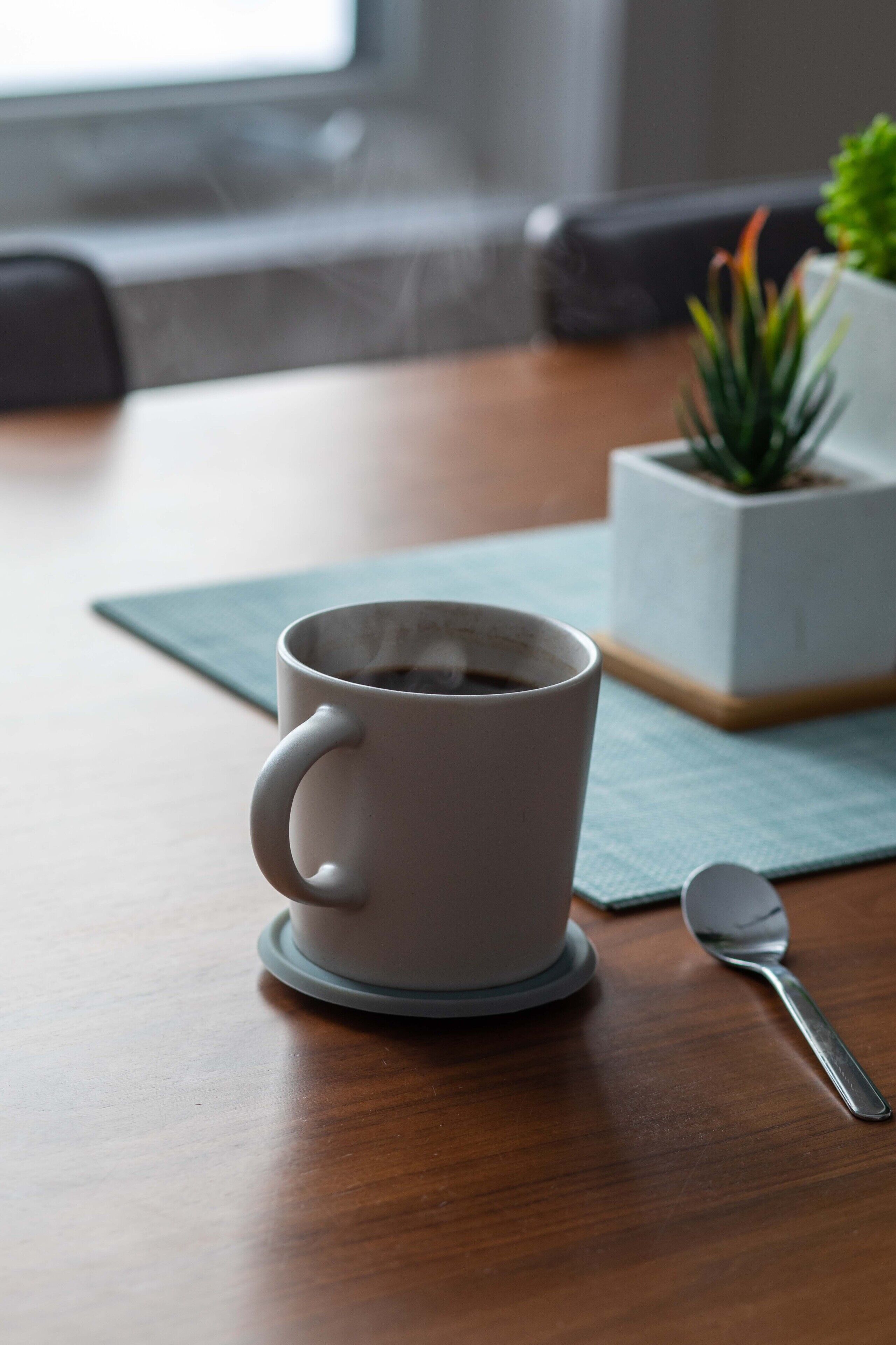 A white cup of coffee on a wooden table with a spoon beside it.