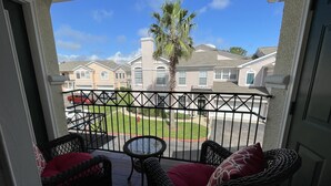 A balcony with wicker furniture and red cushions, overlooking a residential area with houses and a palm tree. - Biloxi border, huge King size Condo (Gulfport)