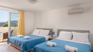 Ett hotellrum med två sängar, båda med blått sänglinne och vita kuddar, ett stort fönster med gula gardiner och en luftkonditioneringsenhet på väggen.