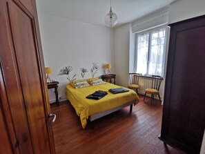 Un dormitorio con suelo de madera, una cama con ropa de cama amarilla, dos mesitas de noche con lámparas, una ventana con cortinas y un radiador.