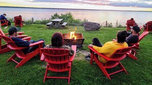 En grupp människor sitter på röda Adirondack-stolar runt en eldstad vid en sjö.
